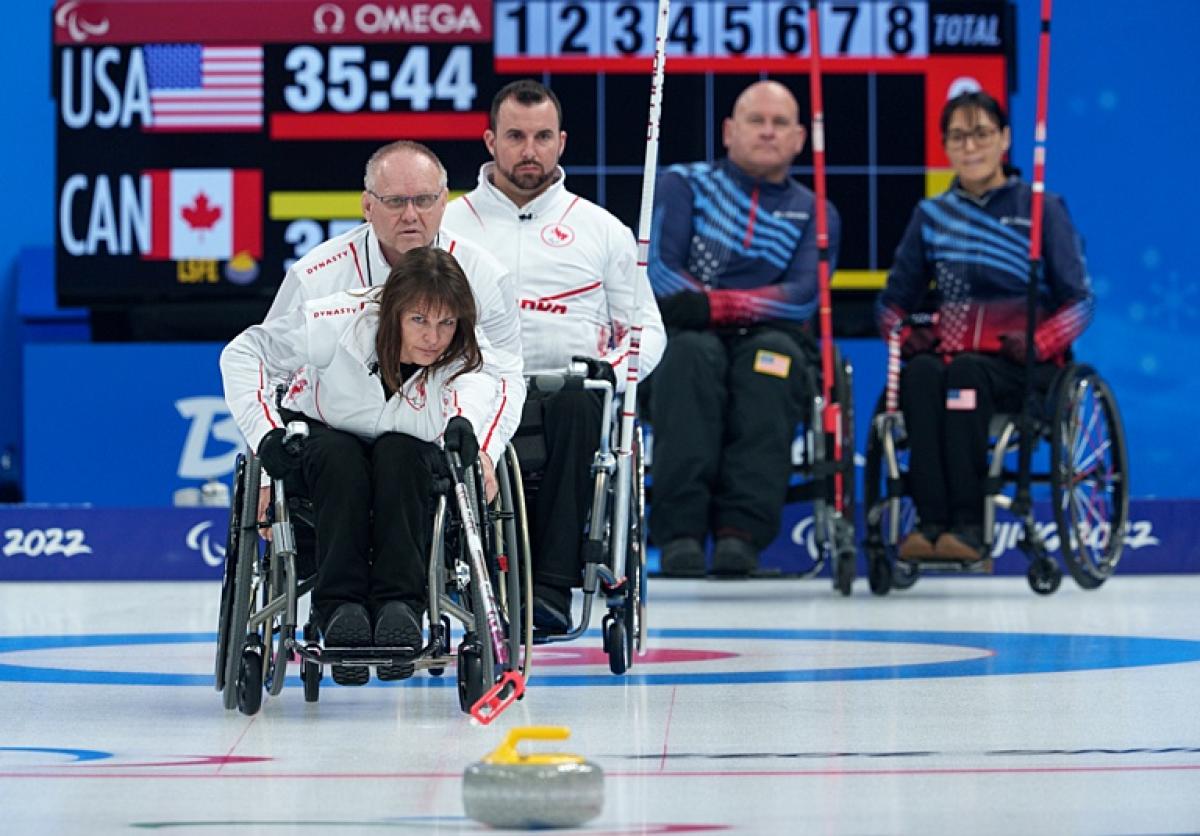 A female wheelchair curling athlete releases a stone with a stick. Four players are watching from behind her